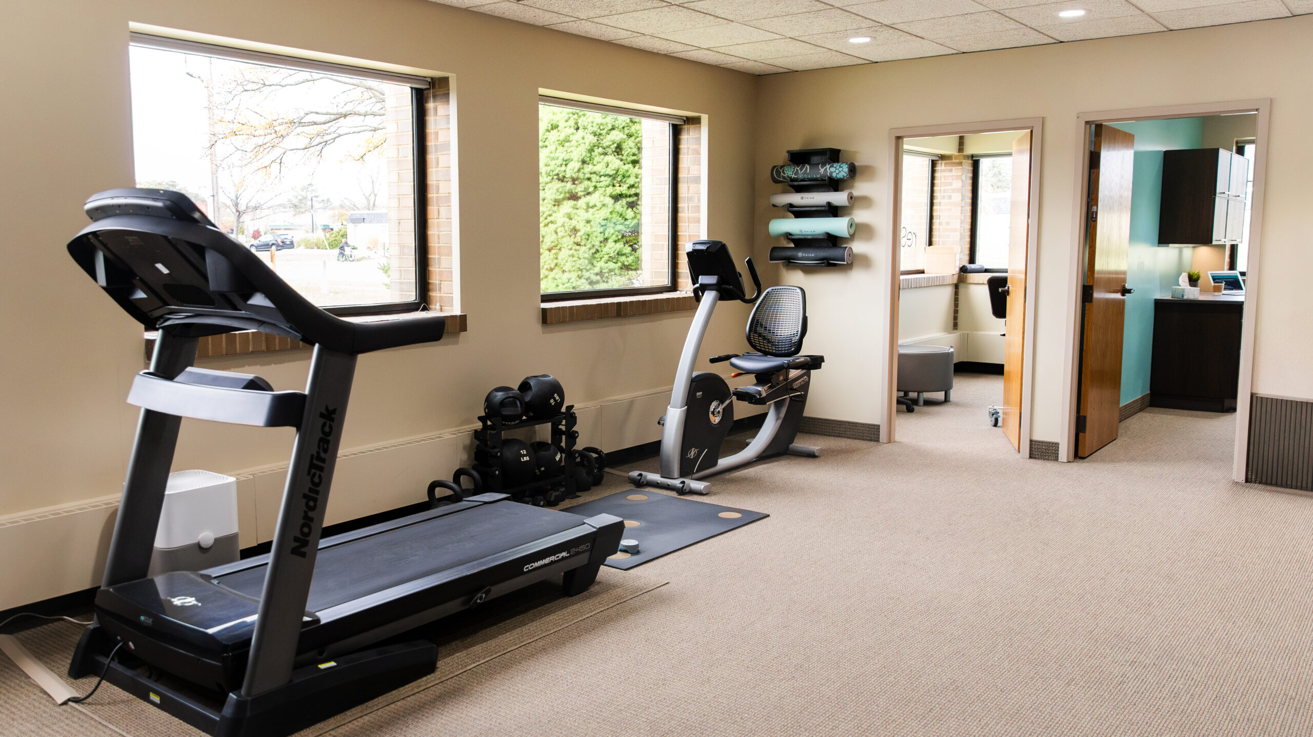 Treadmill and recumbent bike in a physical therapy clinic gym with windows and exercise equipment.